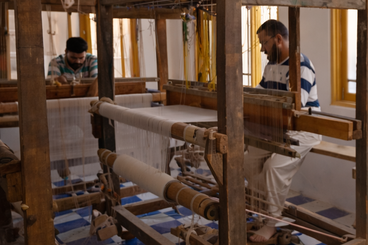 Man using a smartphone in a weaving workshop with looms and windows.