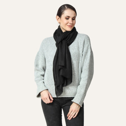 Image of a woman wearing a black cashmere scarf around her neck, knotted in a stylish and elegant way. The knot is positioned off-center and adds a touch of sophistication to the woman's outfit. The woman's expression is confident and composed, with a subtle smile on her face.