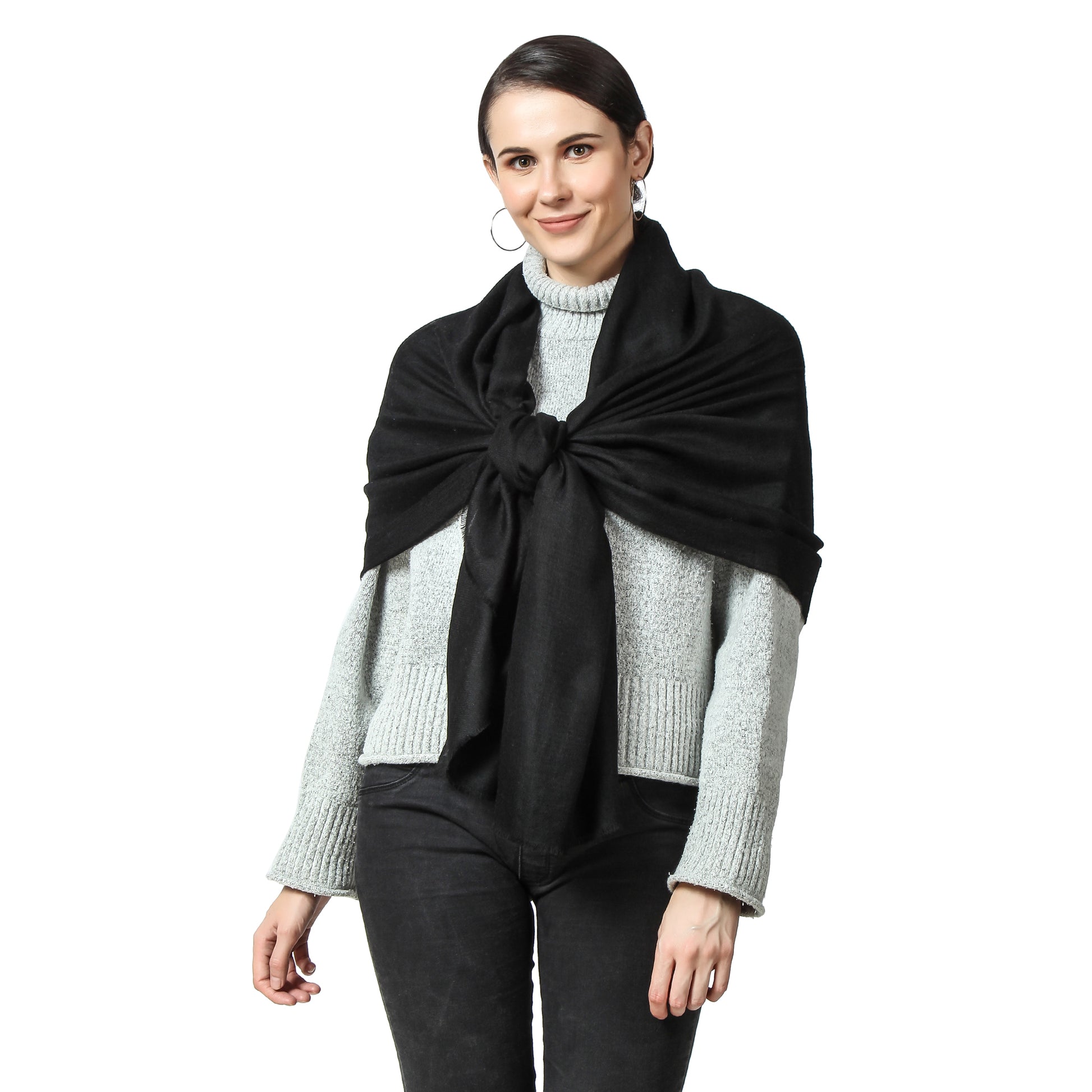 Image of a woman wearing a black cashmere scarf draped over her back and knotted in front. The knot is positioned slightly off-center and adds a stylish touch to the woman's outfit. The scarf is soft and luxurious-looking, and the black color adds to its classic and versatile style. The woman's expression is confident and composed, with a subtle smile on her face.