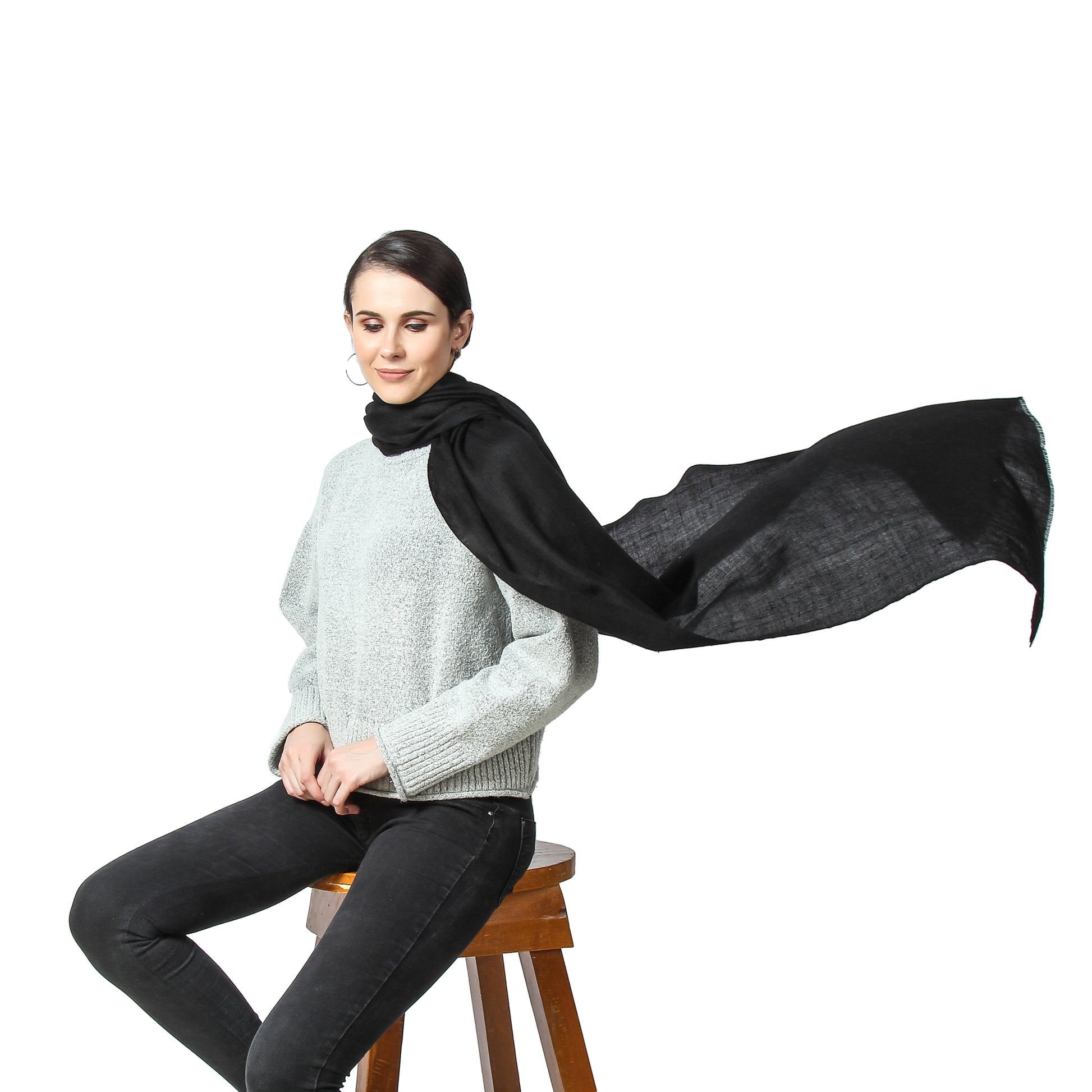Image of a woman sitting on a stool, wearing a black cashmere scarf around her neck. One end of the scarf is blowing in the air, creating a dynamic and elegant effect. The woman's pose is relaxed and confident, with a slight smile on her face.