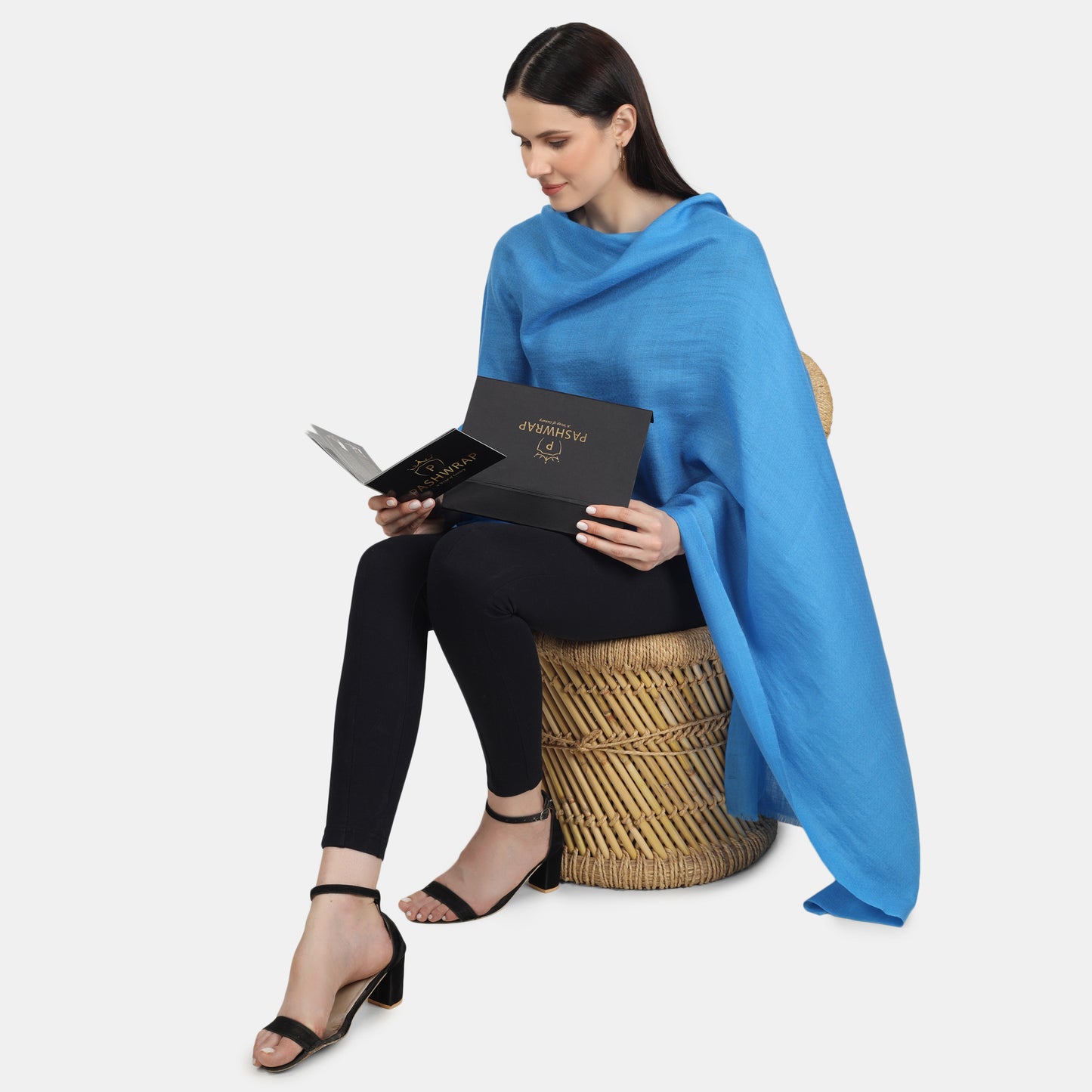 Image of a woman sitting on a chair and wearing a blue cashmere scarf, unboxing a Pashwrap cashmere scarf from a black box. She is holding a product booklet from the Pashwrap brand, which she appears to be reading with interest. The woman's expression is focused and attentive, with a slight smile on her lips. The blue scarf draped around her neck adds a pop of color to her outfit and is in contrast with the black box and the product booklet.