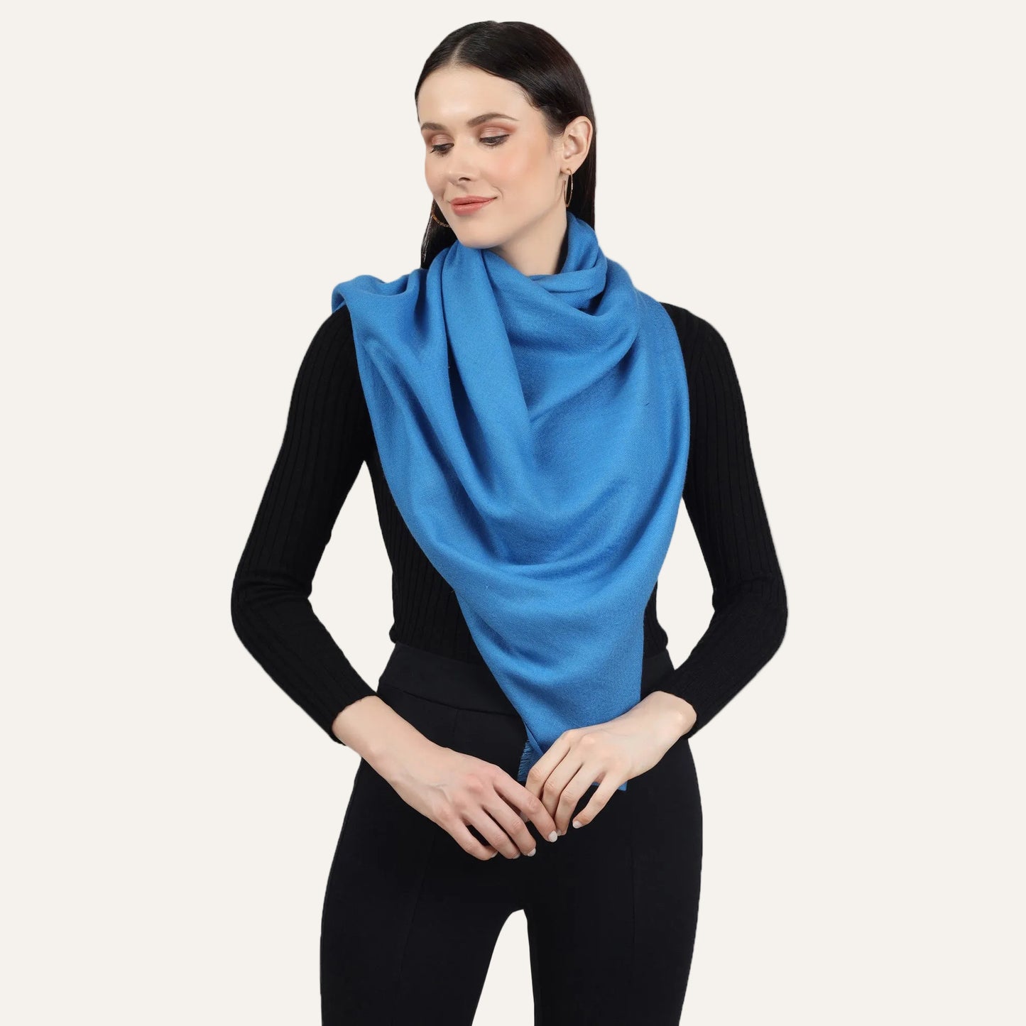 Image of a woman wearing a blue cashmere scarf wrapped around her neck in a stylish and elegant way. The scarf is folded in a neat and precise manner, and the blue color adds a pop of color to the woman's outfit. The scarf appears to be made with high-quality material, and its soft and luxurious texture is visible in the image. The woman's expression is confident and sophisticated, with a subtle smile on her lips.