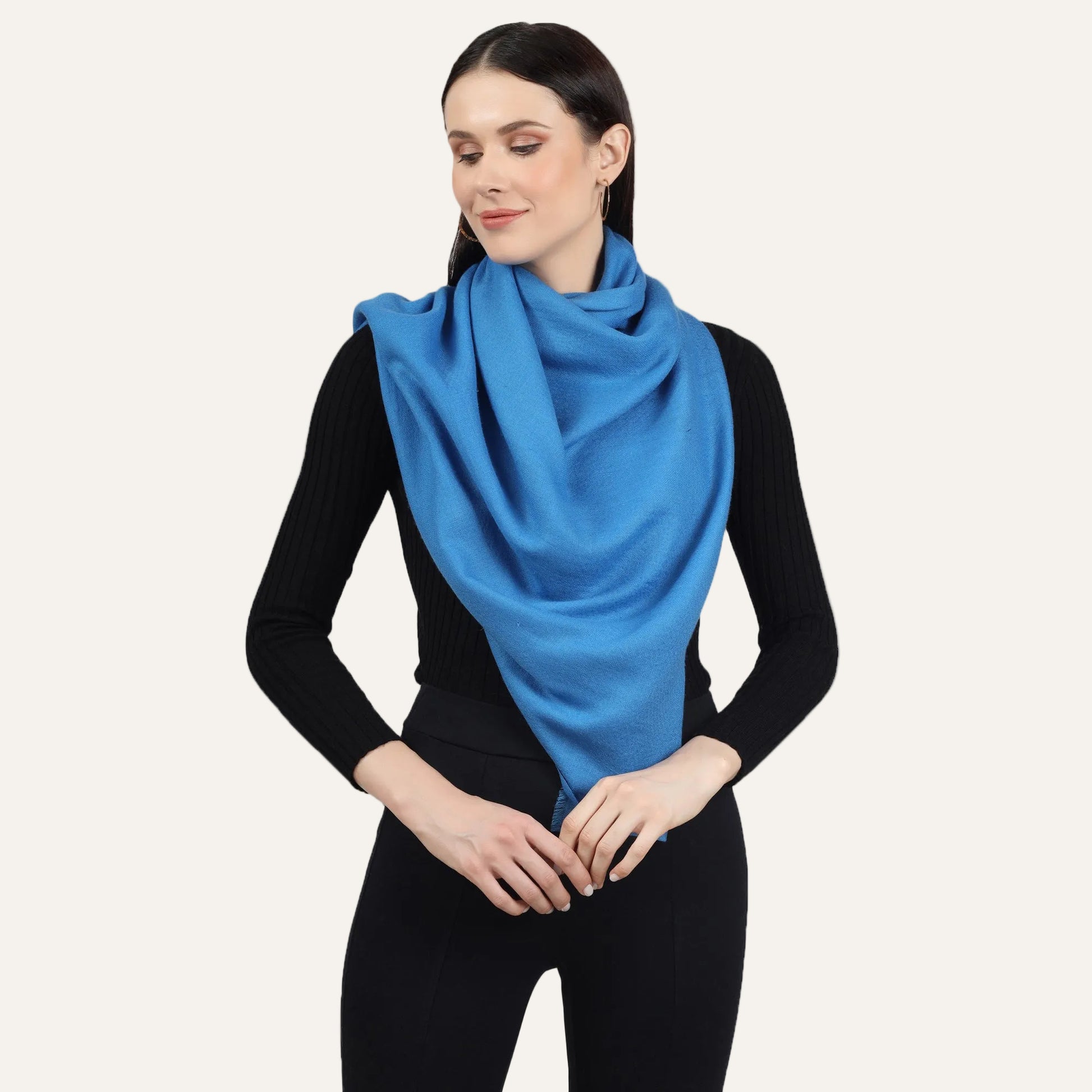 Image of a woman wearing a blue cashmere scarf wrapped around her neck in a stylish and elegant way. The scarf is folded in a neat and precise manner, and the blue color adds a pop of color to the woman's outfit. The scarf appears to be made with high-quality material, and its soft and luxurious texture is visible in the image. The woman's expression is confident and sophisticated, with a subtle smile on her lips.