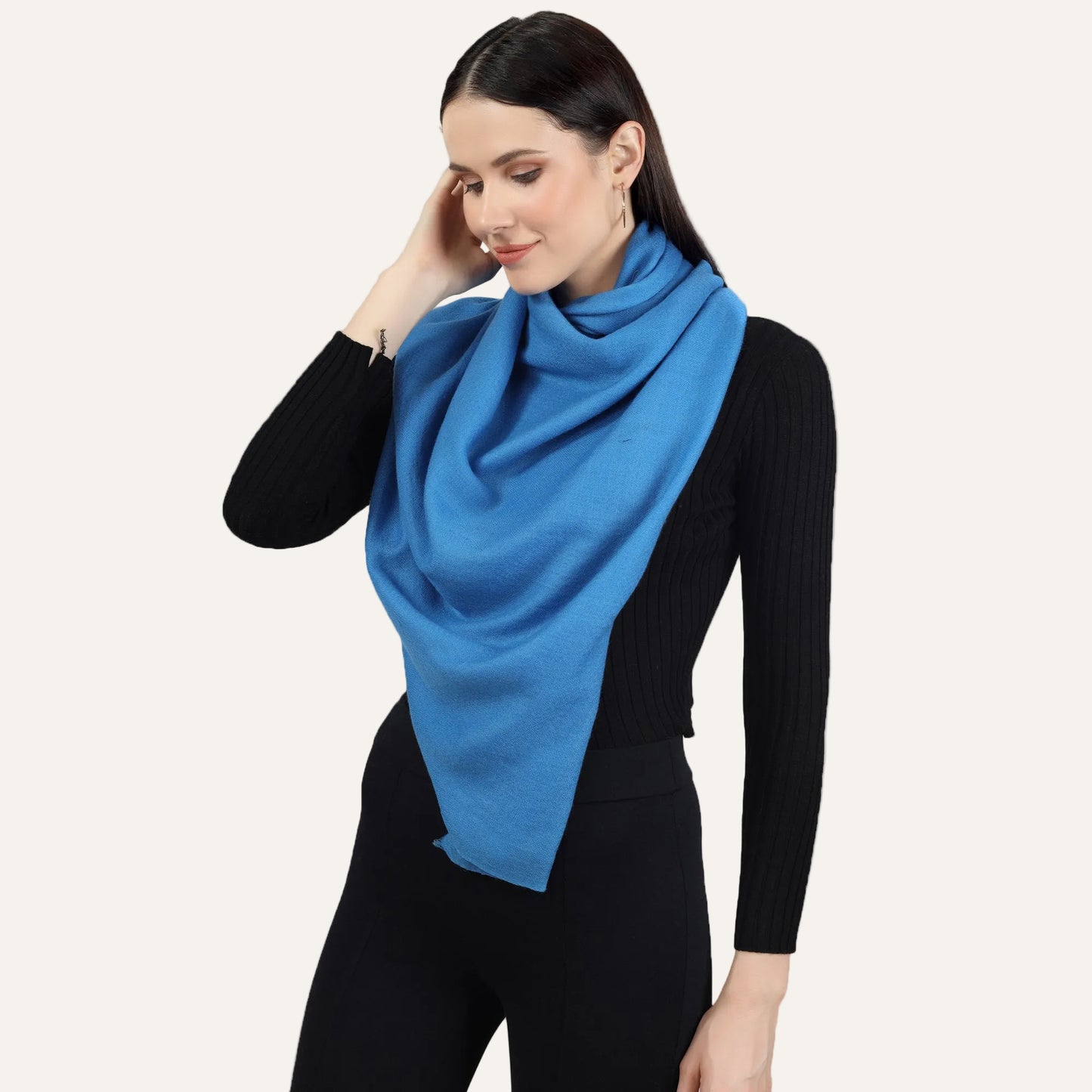Image of a woman wearing a blue cashmere scarf wrapped around her neck in a stylish and elegant way. The scarf is neatly folded and draped around her neck, with one end slightly longer than the other, adding a touch of asymmetry to the look. The blue color adds a pop of color to the woman's outfit, and the soft and luxurious texture of the scarf is visible in the image. The woman's expression is confident and poised, with a subtle smile on her lips.
